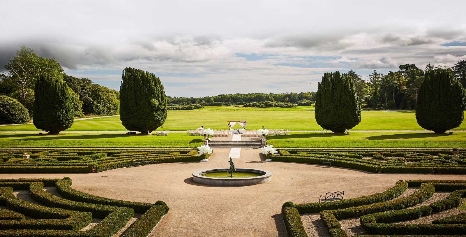 a fountain in a circular area with hedges and trees