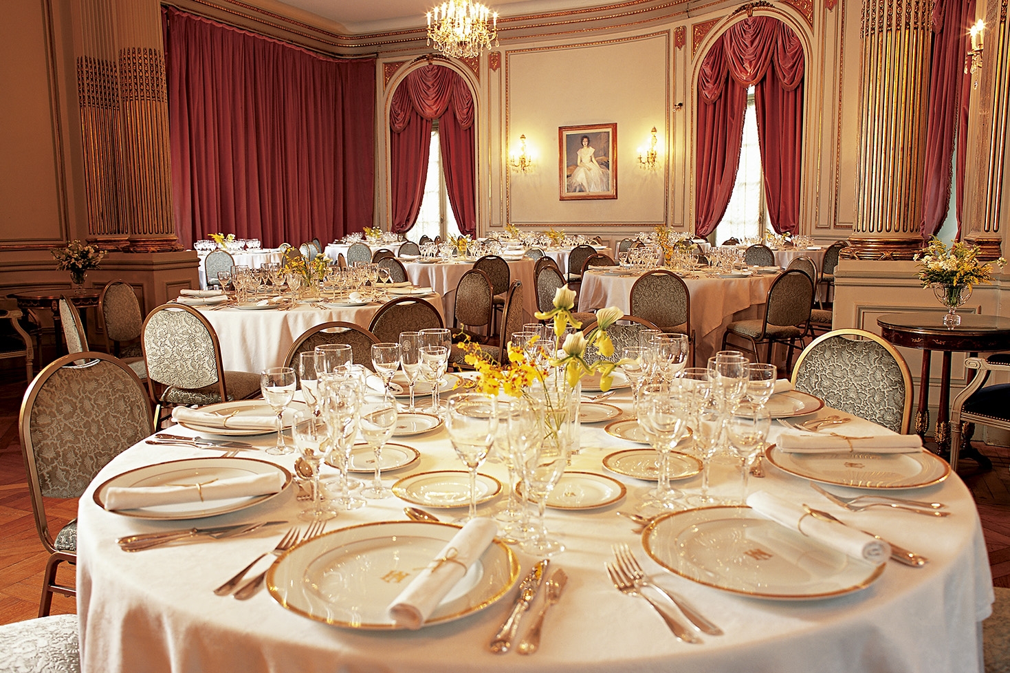 A grand, formal banquet room with high ceilings, gold-accented columns, dusty-rose velvet curtains, and round dining tables meticulously set with fine china, crystal glassware, and yellow floral centerpieces.