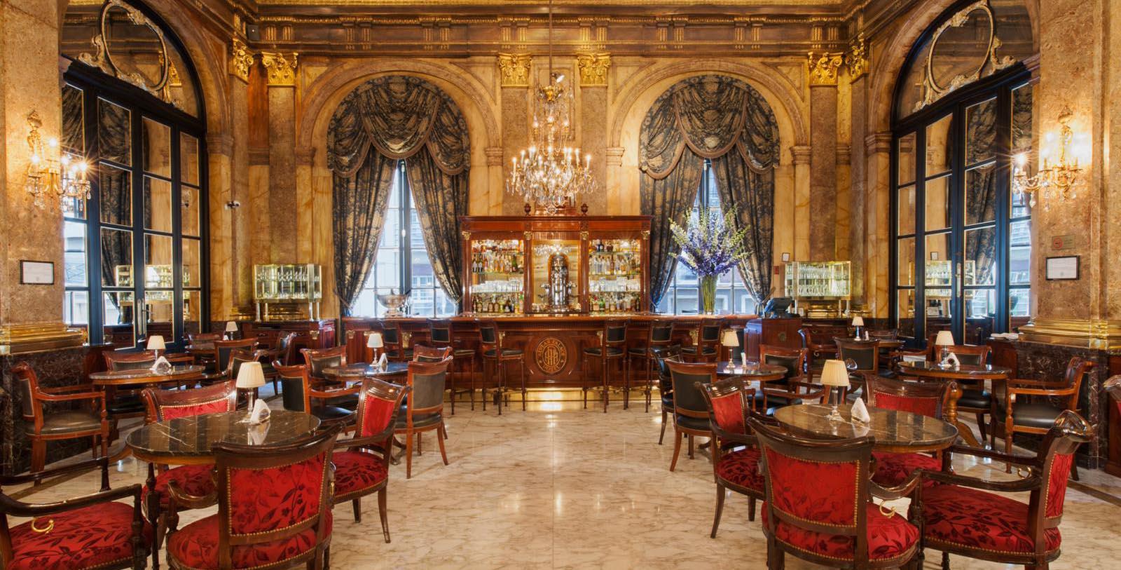 An opulent hotel bar and lounge with marble walls, large arched mirrors, rich red upholstered chairs, polished wooden tables, and a central back bar framed by heavy drapes and a crystal chandelier.