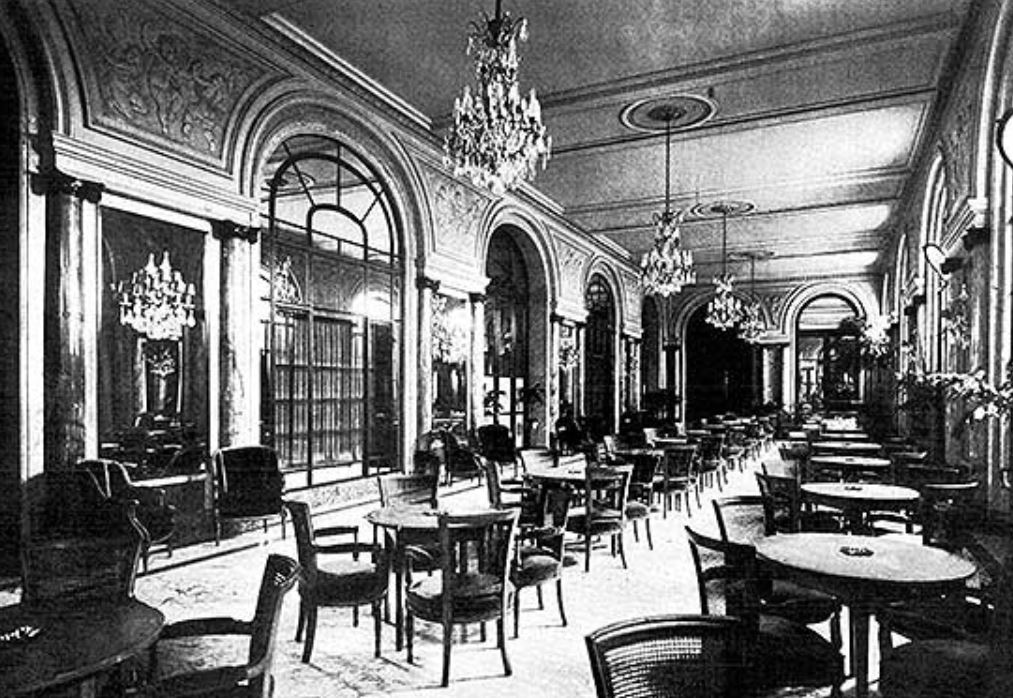 A historic black-and-white photograph of a grand hotel dining hall or lounge, lined with tall arched French doors, ornate wall moldings, marble columns, hanging crystal chandeliers, and scattered dining tables.