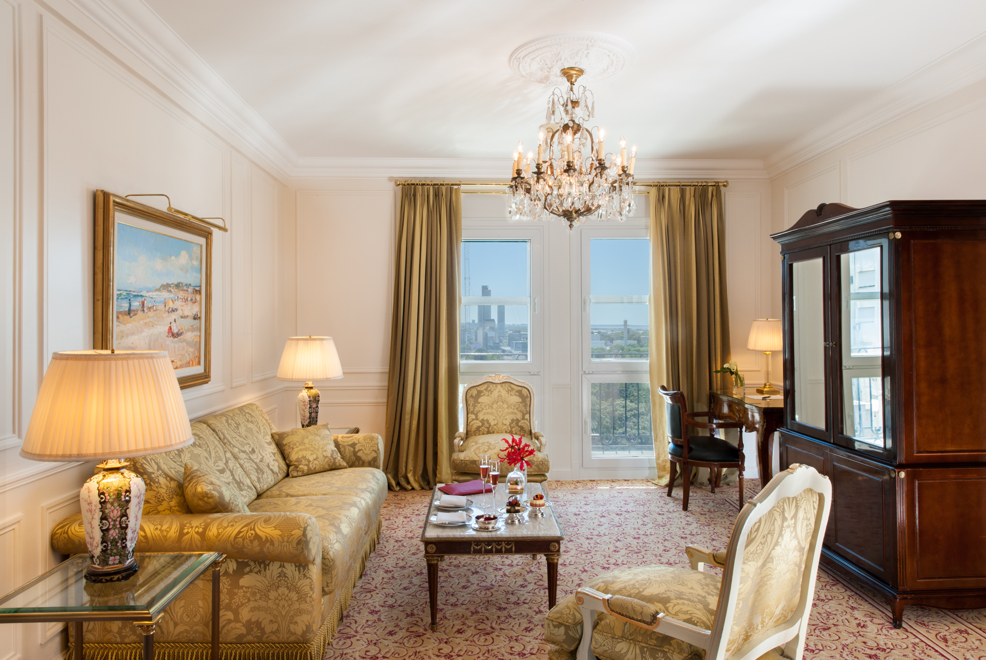 A bright luxury hotel living room featuring gold damask upholstered seating, a crystal chandelier, dark wooden furniture including a large armoire, and tall windows with gold drapes offering a city view.