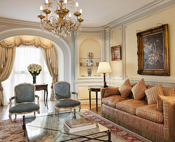 An elegantly decorated luxury suite sitting room featuring classical European decor, a crystal chandelier, a striped sofa, plush blue armchairs, framed oil paintings, and an arched window nook with draped curtains.
