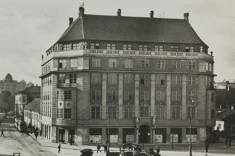 A historical black-and-white photograph depicting the exterior facade of a grand multi-story urban building, showing streetcars and pedestrians on the road below.
