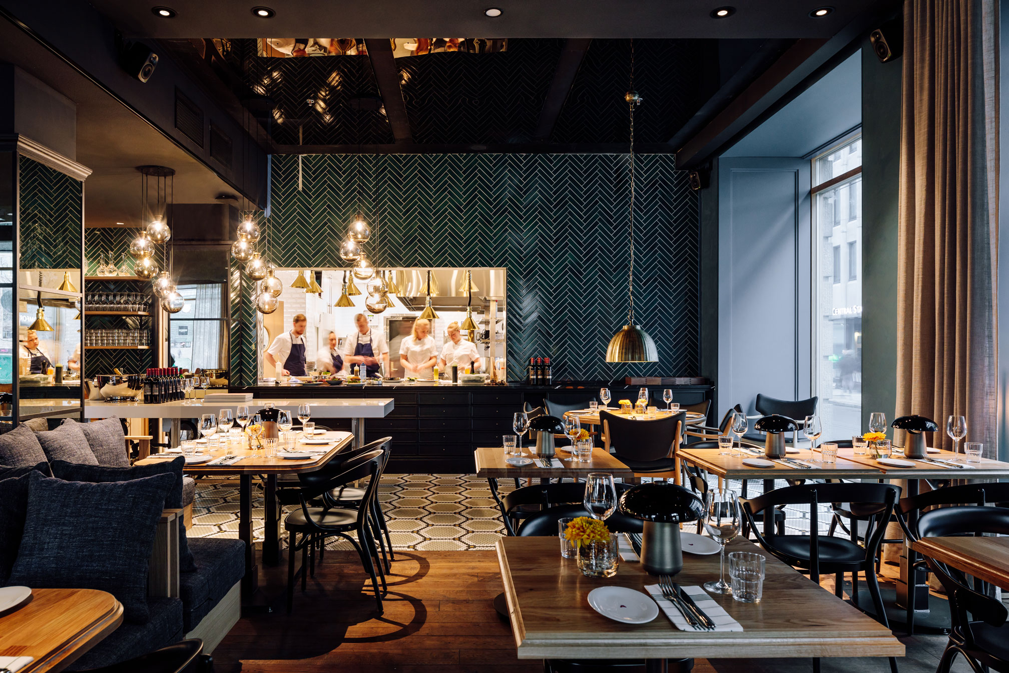 A modern, upscale hotel restaurant featuring dark wood tables, black chairs, a herringbone dark green tiled wall behind an open kitchen pass, and glowing pendant lights.