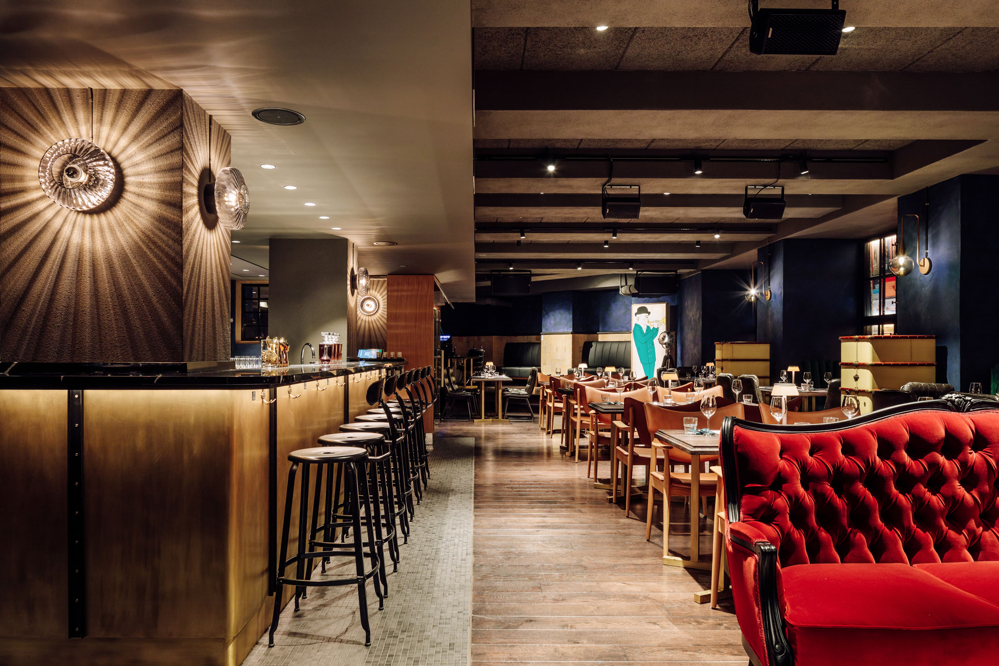 A stylish, dimly lit hotel bar and lounge featuring a long illuminated metallic bar counter with tall stools, dining tables, dark blue walls, and a prominent red tufted velvet sofa in the foreground.