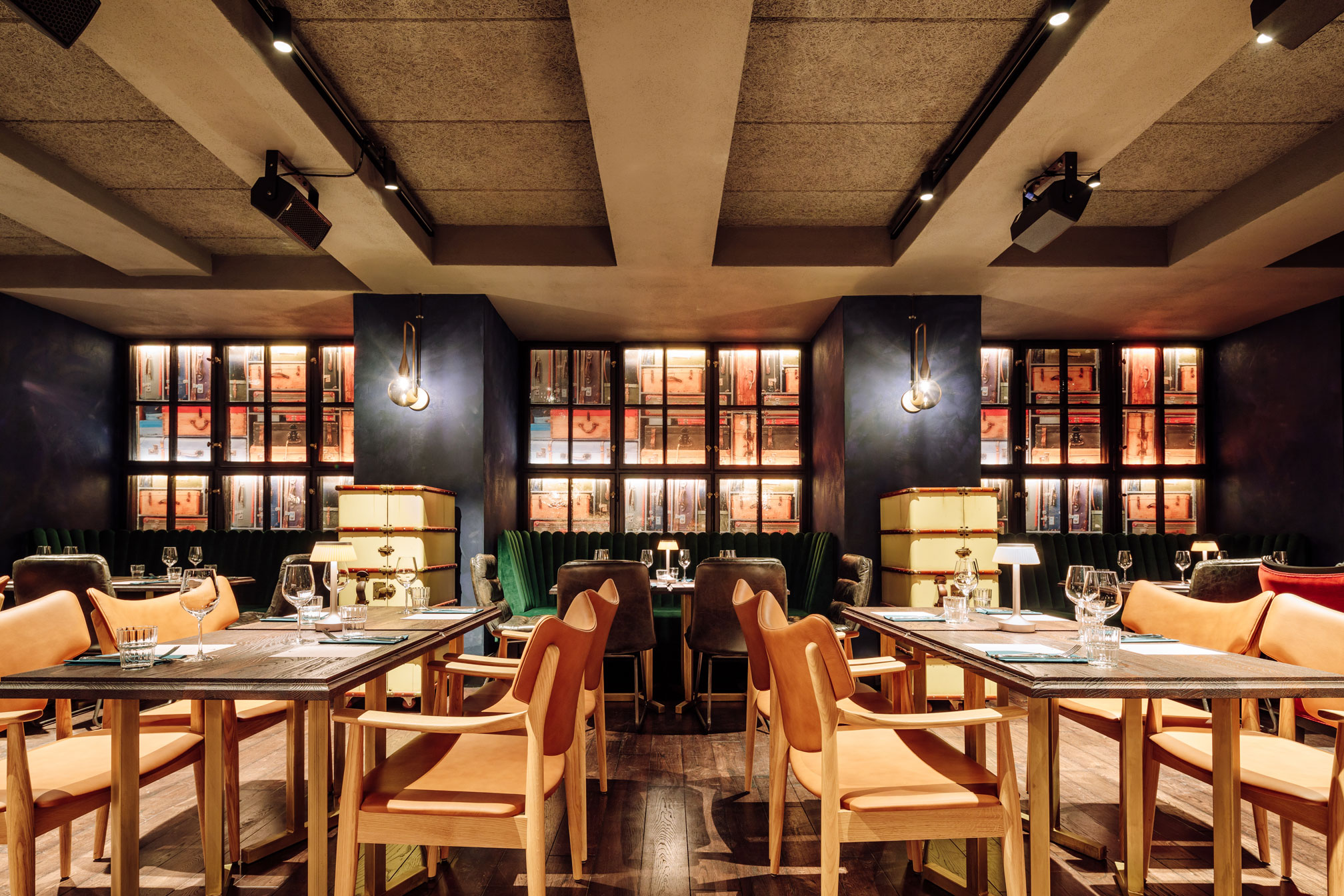 A moody, upscale restaurant dining area with light wood tables, tan leather armchairs, dark teal velvet booth seating, and back-lit glass display cabinets filled with vintage luggage.