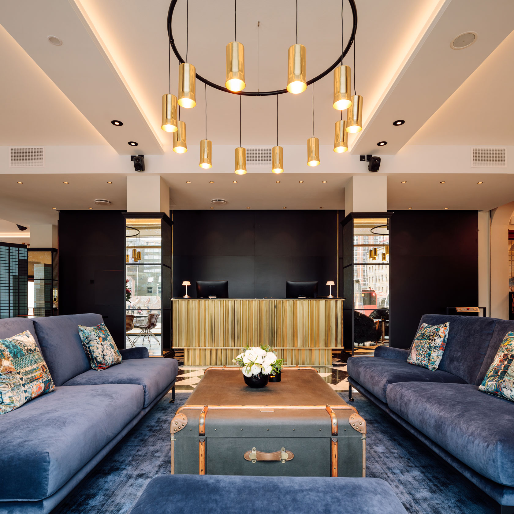 A modern hotel lobby featuring blue velvet sofas arranged around a dark trunk coffee table, situated in front of a ribbed brass reception desk, with a circular brass pendant chandelier hanging overhead.