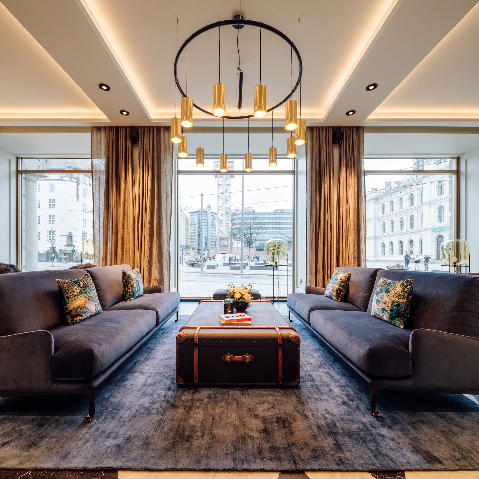 A stylish hotel sitting area with facing dark grey plush sofas, a trunk coffee table in the center, a large round chandelier with gold cylindrical lamps, and large floor-to-ceiling windows with bronze drapes looking out onto a city street.