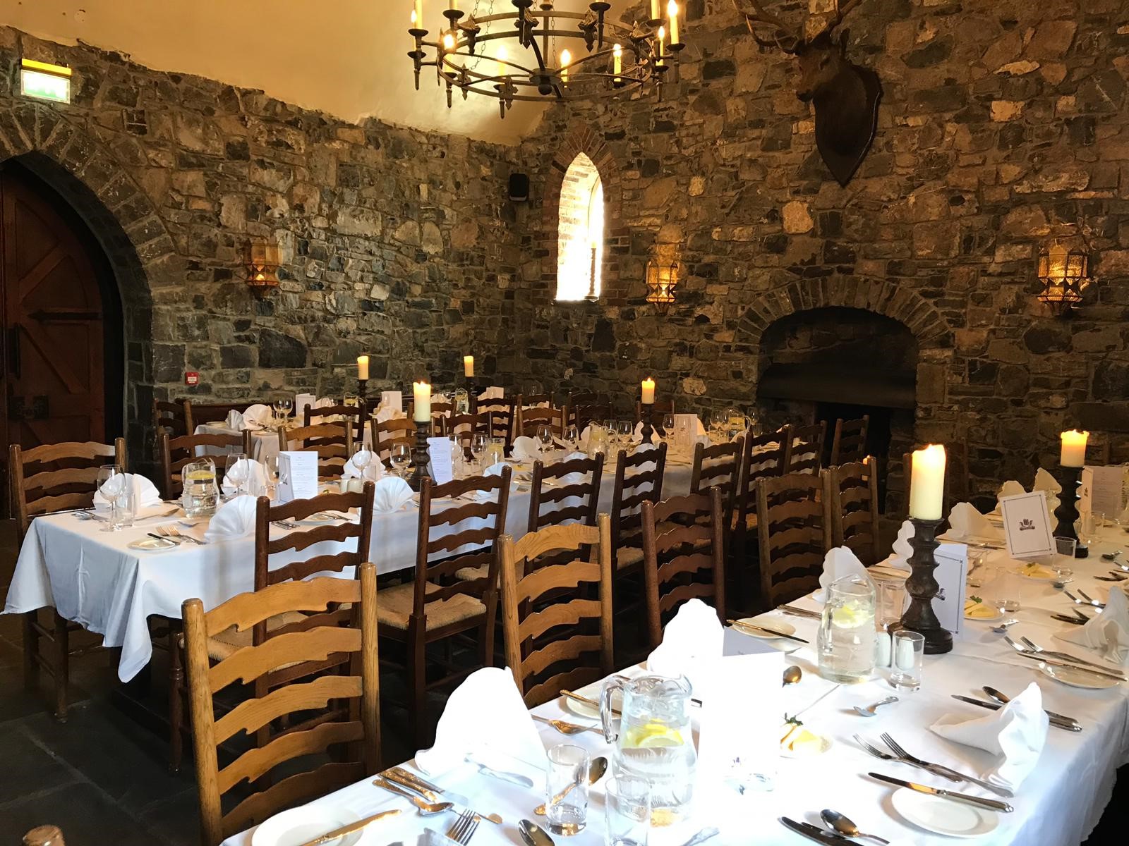 A rustic castle dining hall featuring exposed stone walls, a large fireplace, high-backed wooden chairs around long white-clothed tables, candle centerpieces, and a rounded iron chandelier.