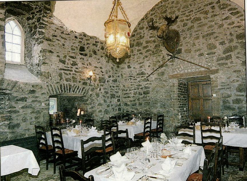 A historic dining room set in a stone castle chamber with high vaulted ceilings, featuring tables set with white linens, wooden ladder-back chairs, a large hanging lantern light fixture, and a mounted animal head on the stone wall.