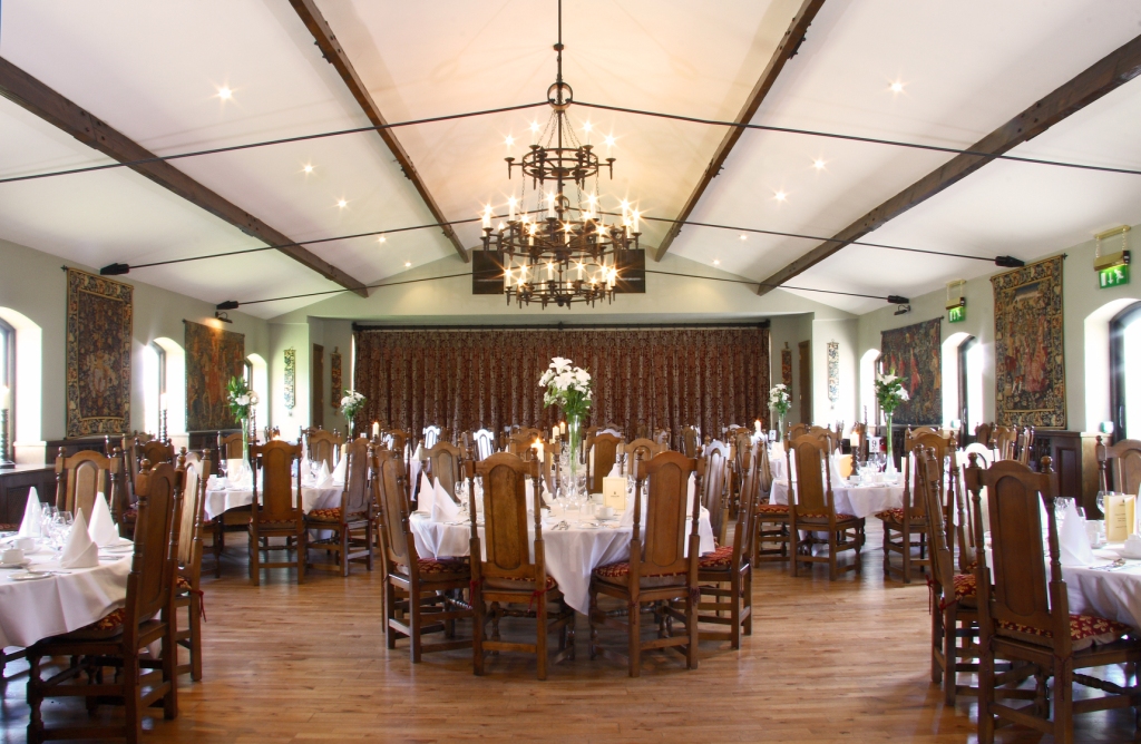 A grand banquet hall with high vaulted ceilings, exposed wooden beams, a large wrought-iron chandelier, hardwood floors, and round tables set with white linens and ornate high-backed wooden chairs.