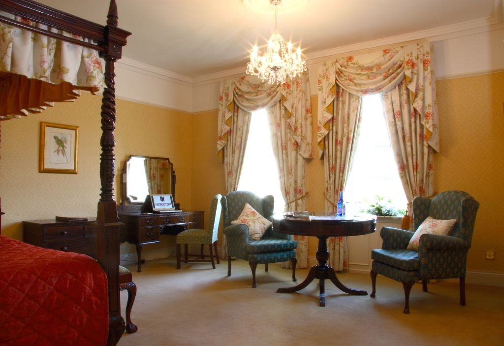 A traditional hotel suite decorated in warm yellow tones, featuring a four-poster dark wood bed, floral drapes, a crystal chandelier, two patterned armchairs around a round wooden table, and a vanity desk.