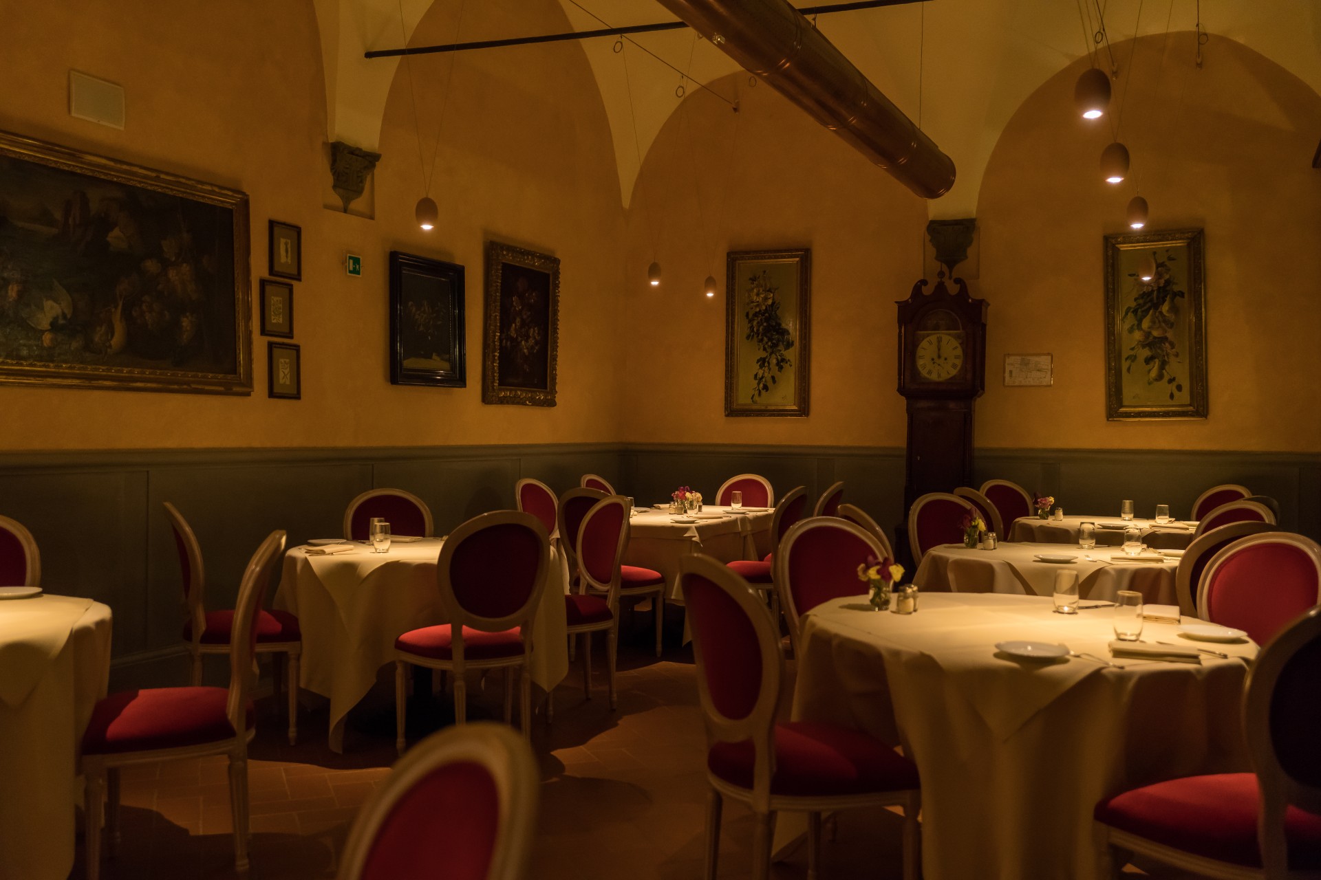 An intimate, softly lit hotel dining cellar under vaulted plaster ceilings, featuring white-clothed tables surrounded by rich red oval-backed chairs, framed art on plaster walls, and a classic grandfather clock in the corner.