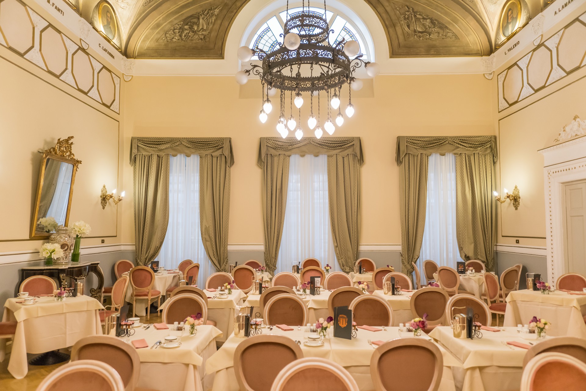A elegant hotel dining hall set with white-clothed tables and dusty rose oval-backed chairs, featuring high arched windows framed by olive green drapes, a vaulted frescoed ceiling, and a large black wrought-iron chandelier