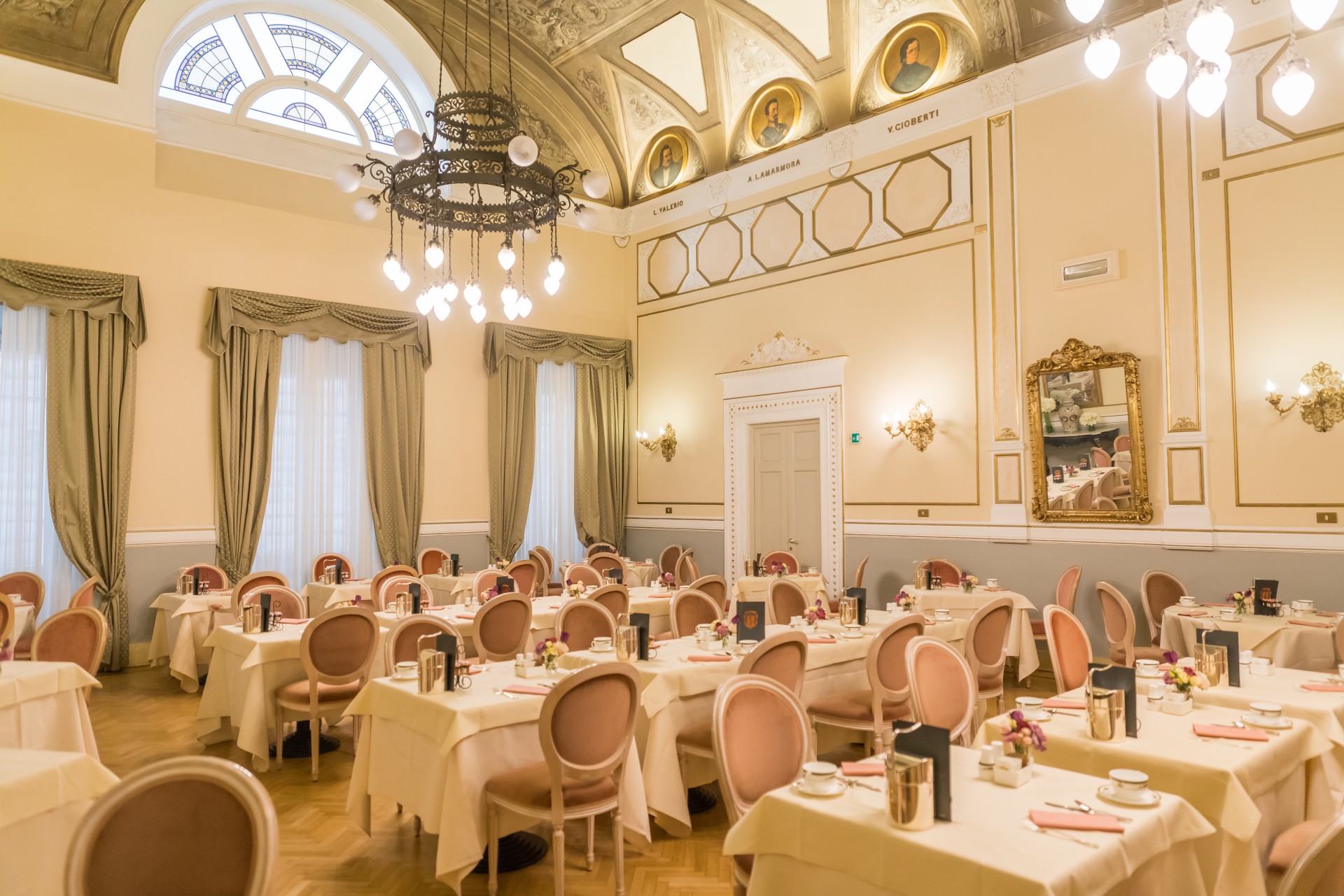 A wide view of a grand historic dining hall featuring white-clothed tables with dusty rose oval-backed chairs, herringbone parquet floors, a vaulted frescoed ceiling with portrait medallions, and an ornate black iron chandelier.