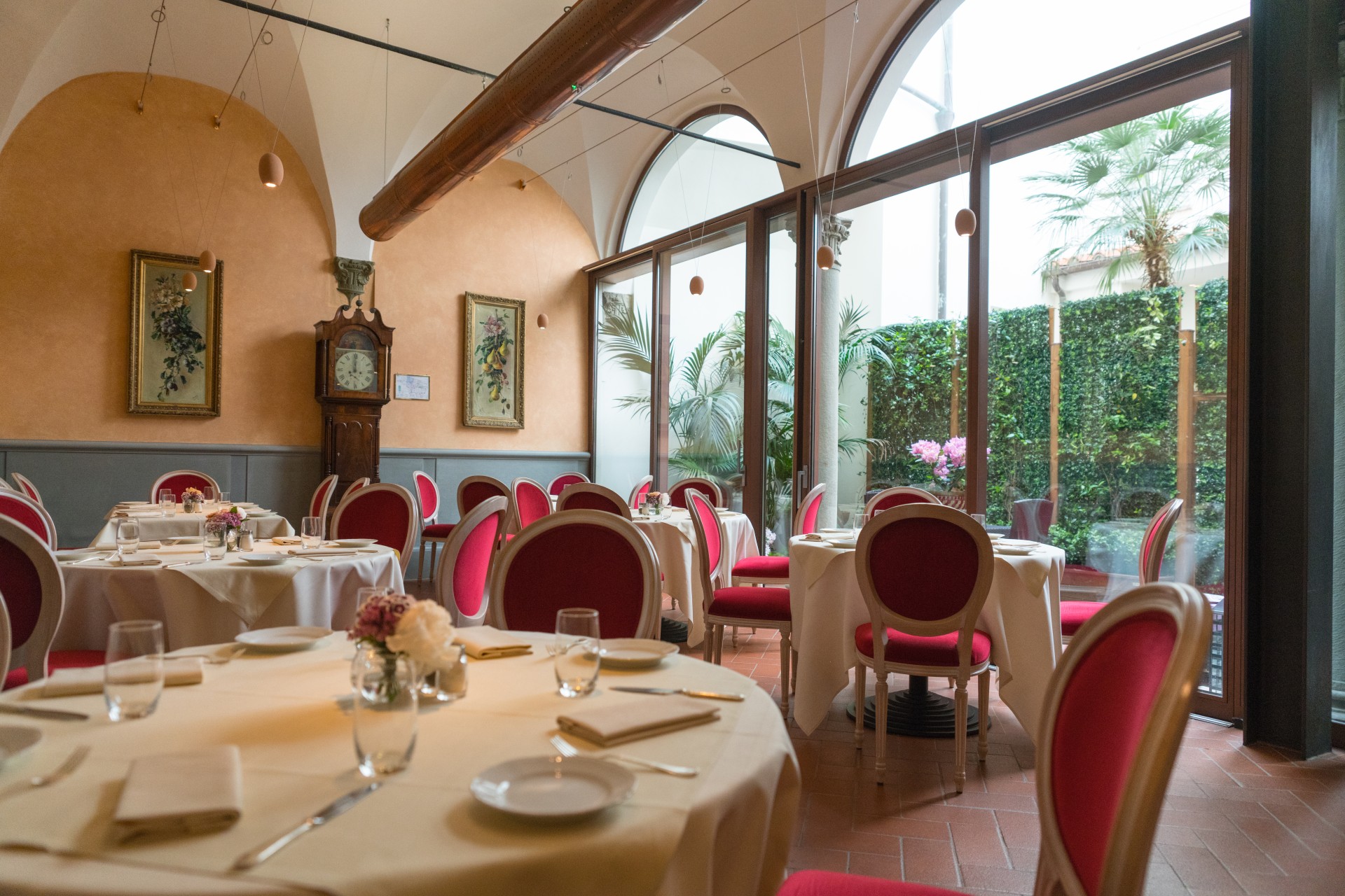 A bright hotel dining room with terracotta floor tiles, vaulted plaster ceilings, round tables in white linens with red velvet-trimmed oval-back chairs, framed artwork, a grandfather clock, and large arched glass windows looking out onto a lush green cour