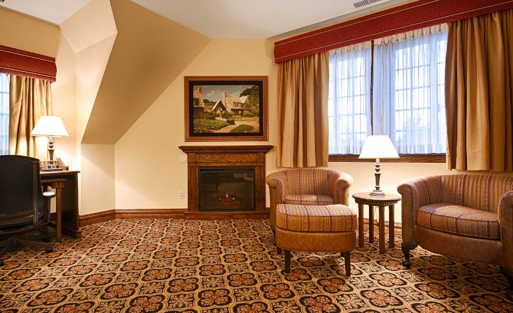 Cozy hotel sitting area featuring a wooden fireplace, framed artwork above the mantel, plush striped armchairs with a matching ottoman, patterned carpet, a desk with a office chair, and warm curtains over multi-pane windows.