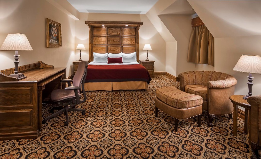 Warmly lit hotel suite featuring a king-size bed with a large wooden panelling headboard and red bed throw, a wooden writing desk with an office chair, a comfortable armchair with matching ottoman, patterned carpet, and bedside lamps.
