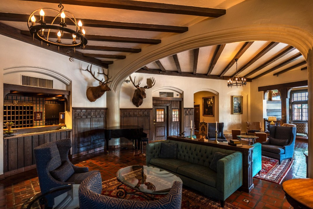 Rustic lodge lounge featuring exposed ceiling beams, wrought-iron chandeliers, mounted elk and moose heads on wood-paneled walls, a grand piano, and comfortable seating including a teal tufted sofa and patterned wingback armchairs.