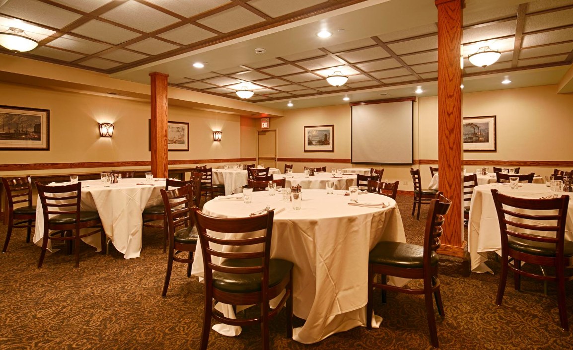 A banquet room set up with round tables draped in white tablecloths and paired with dark wooden chairs, featuring wood support columns, grid coffered ceiling, wall sconces, framed artwork, and a pull-down projection screen at the far wall.