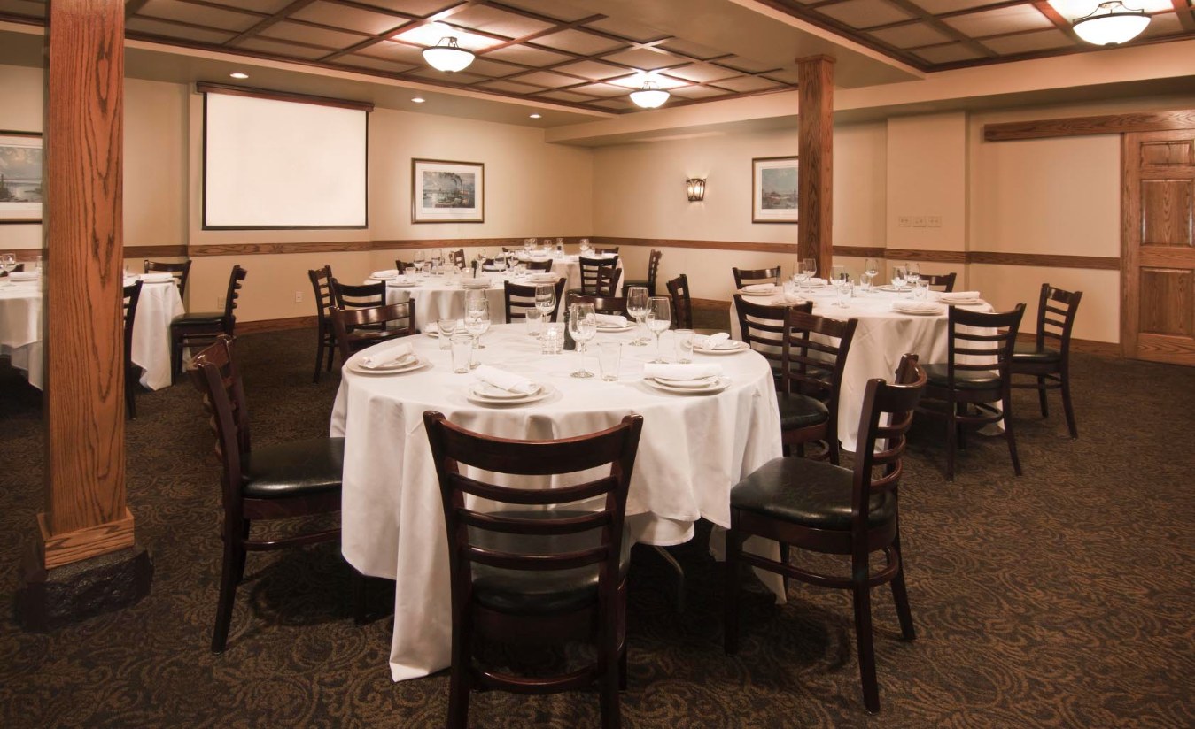 Banquet dining room filled with round tables covered in white tablecloths, set with place settings and drinking glasses, surrounded by dark wooden chairs under a coffered ceiling with light fixtures and wooden support pillars.