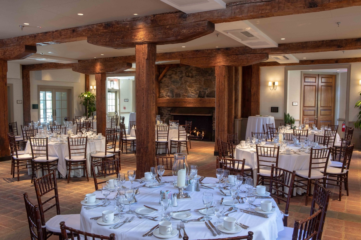 A rustic dining hall or event room with exposed wooden beams and pillars, round tables set with white tablecloths and formal place settings, Chiavari chairs, and a stone fireplace in the background.