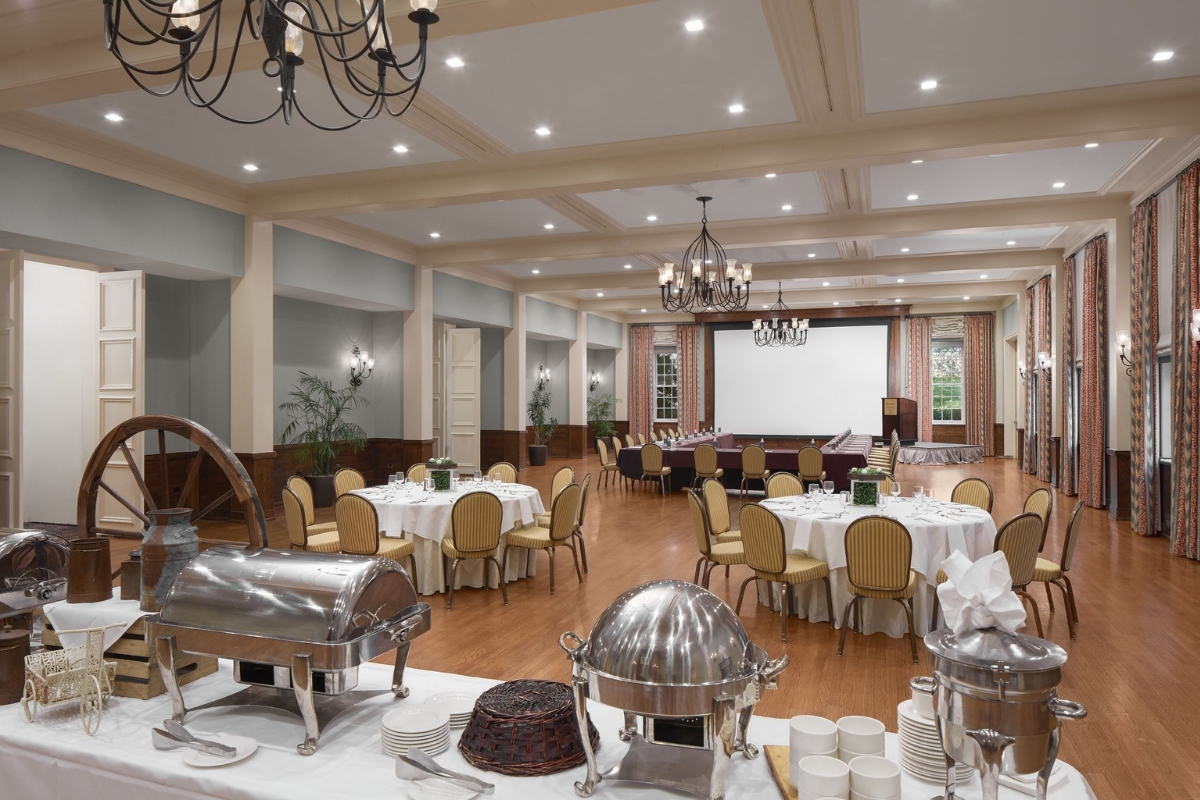 A large event hall featuring a buffet setup in the foreground with chafing dishes, round banquet tables with gold chairs, light blue walls, wrought-iron chandeliers, and a projection screen with a U-shaped conference table setup at the far end.