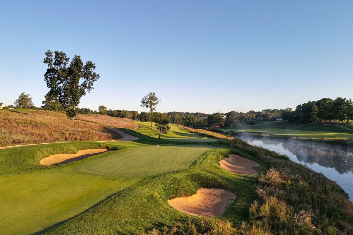 A scenic golf course green featuring sand traps, a pin flag, a calm body of water with early morning mist on the right, and rolling grassy hills under a clear sky.