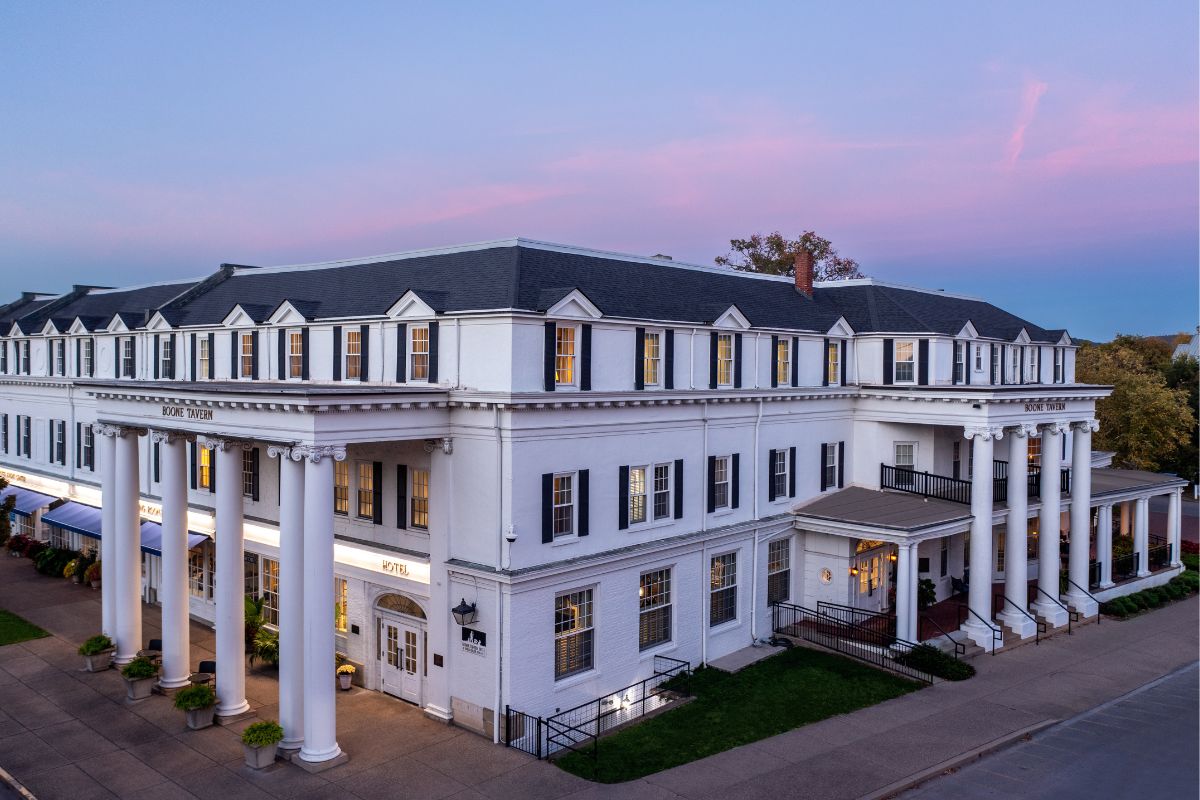An exterior twilight view of the historic Boone Tavern Hotel featuring white brick walls, dark shutters, classic grand white columns supporting prominent covered entrance porticos, and a pink-and-purple dusk sky overhead.