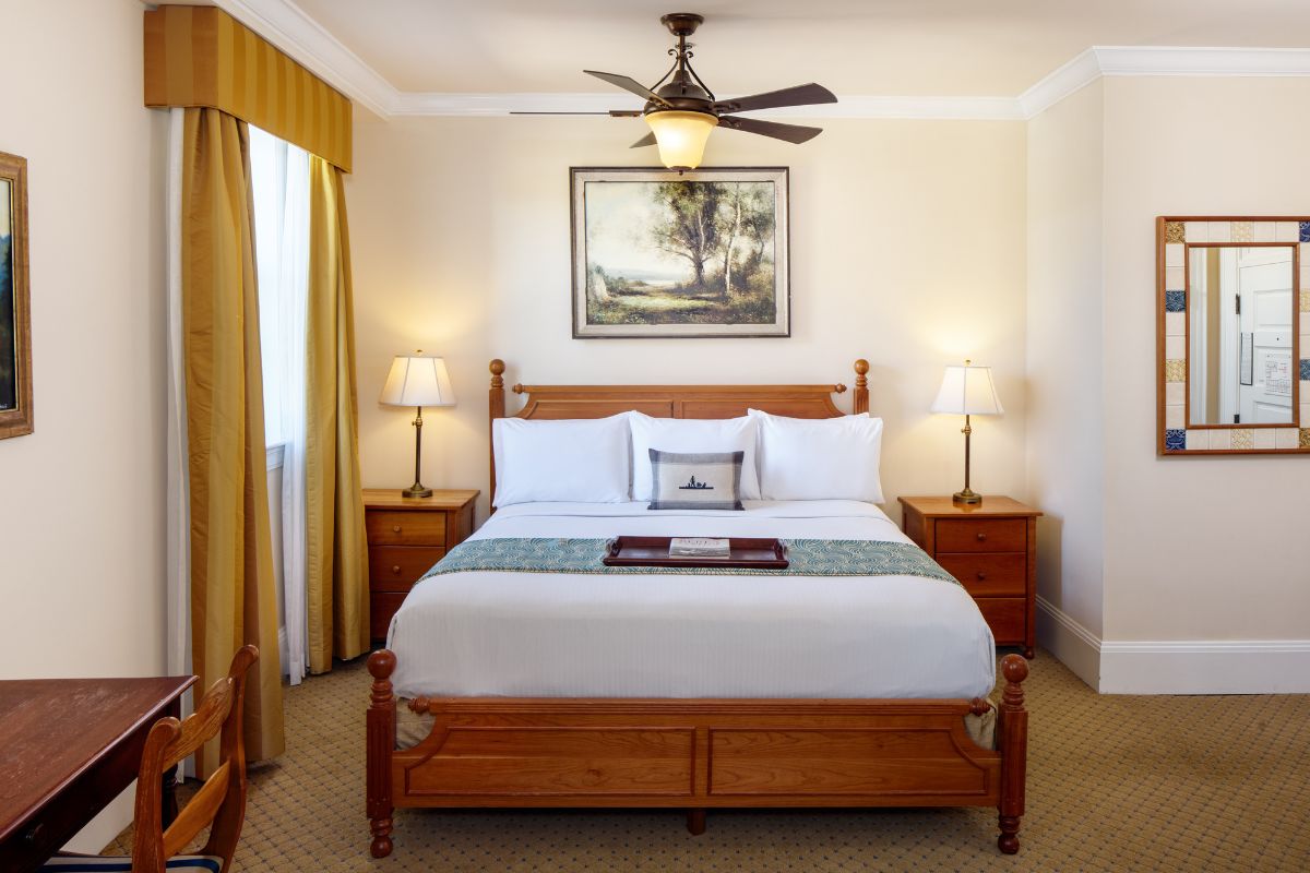 A cozy, traditional hotel bedroom featuring a warm wooden bed frame with white linens, wooden nightstands with bedside lamps, a ceiling fan, landscape artwork above the bed, and gold drapes covering the window.
