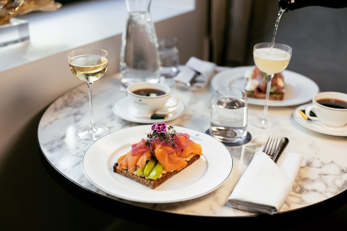 An elegant table arrangement with a smoked salmon and asparagus open-faced sandwich, glasses of sparkling wine being poured, coffee cups, and water glasses on a round marble tabletop.