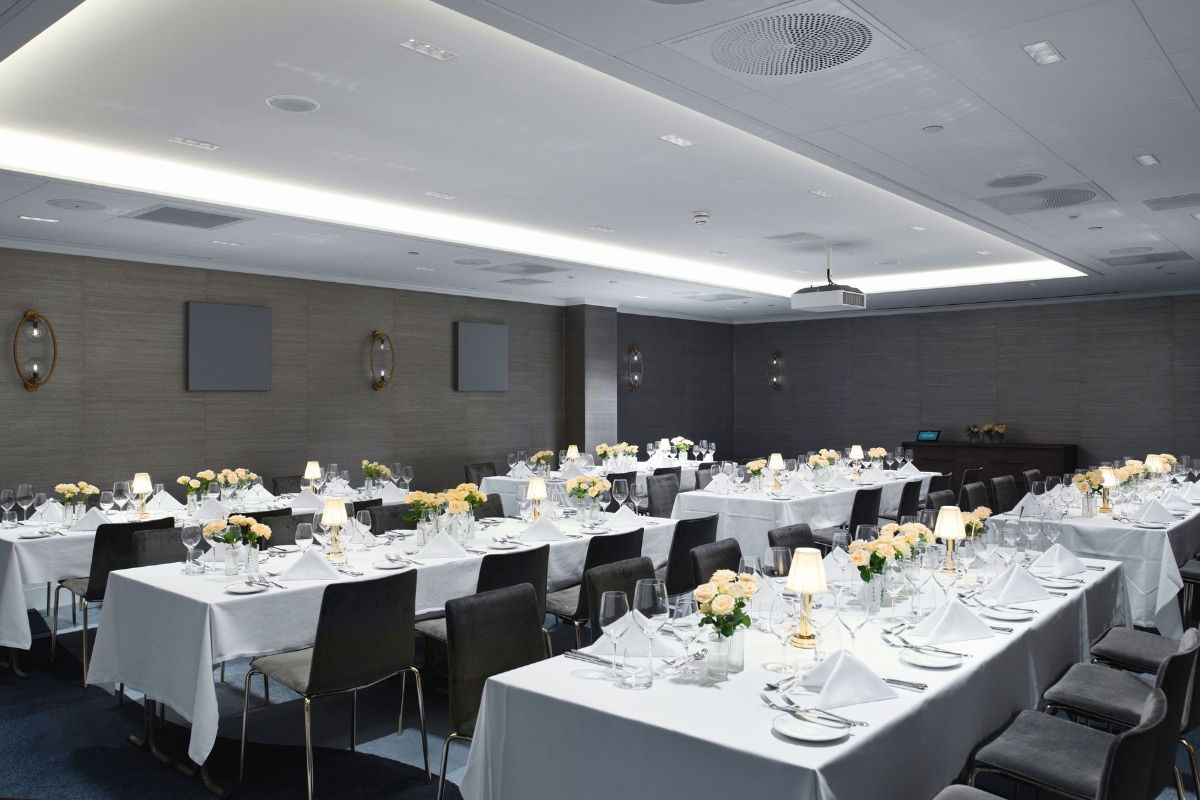 A modern, formal banquet or event room set up with long rectangular tables covered in white tablecloths, dark chairs, small floral centerpieces, table lamps, and ceiling-mounted projector lighting.