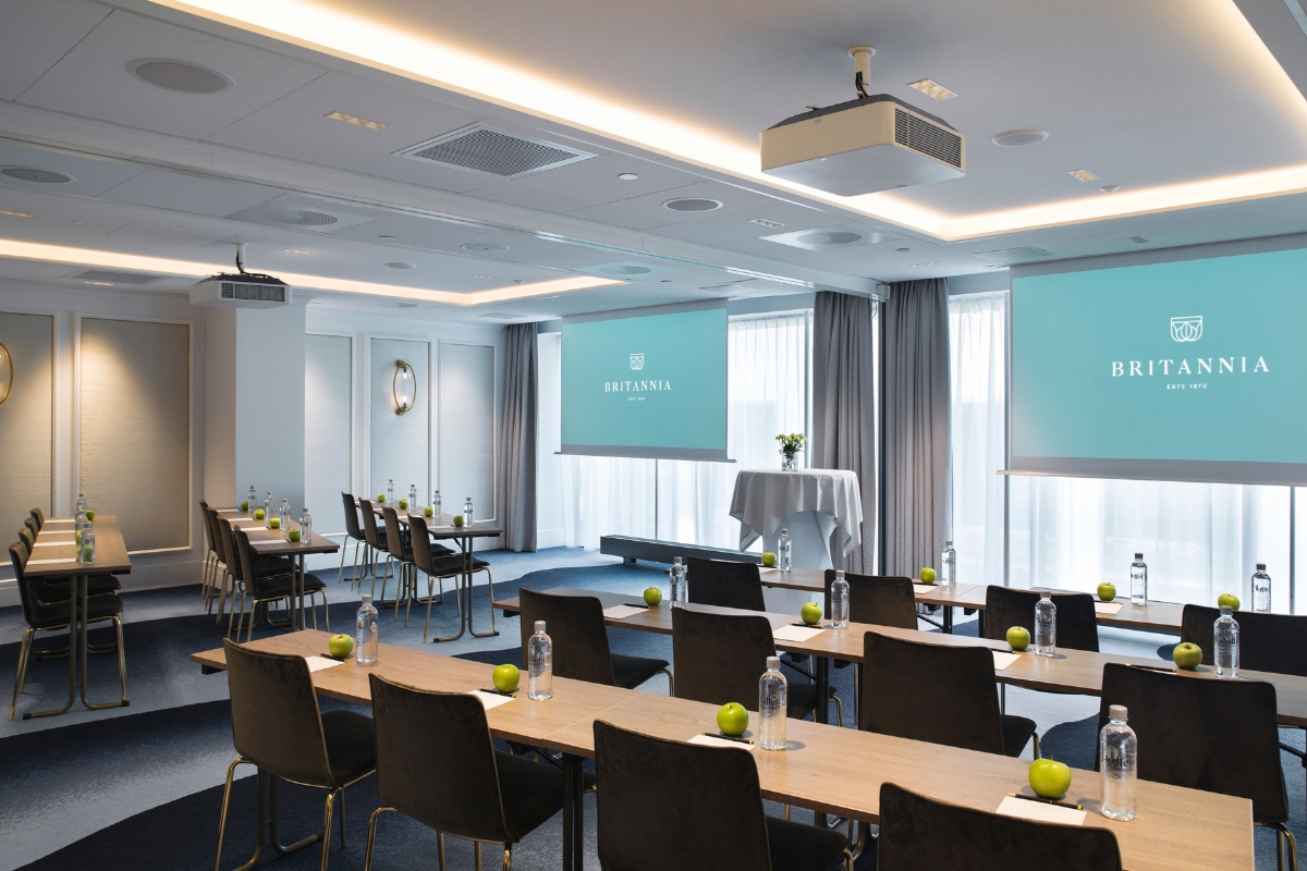 A modern presentation or conference room set up classroom-style with wooden desks, dark chairs, bottled water, green apples, and two large projection screens displaying the Britannia logo.