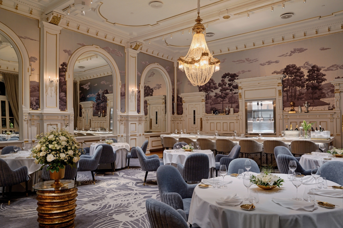 A grand, opulent dining hall with high ceilings, a massive central crystal chandelier, landscape wallpaper murals, round dining tables set with white tablecloths, and dusty blue upholstered chairs.