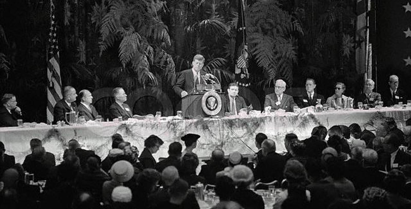 a man standing at a podium with a group of people sitting at a table