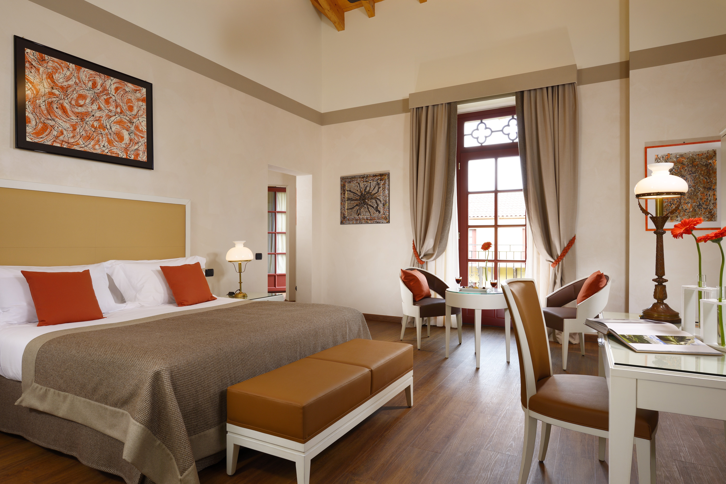 A spacious hotel suite featuring high ceilings, a king bed with orange accent pillows and a leather bench, a small dining table near a balcony door, and a white writing desk.