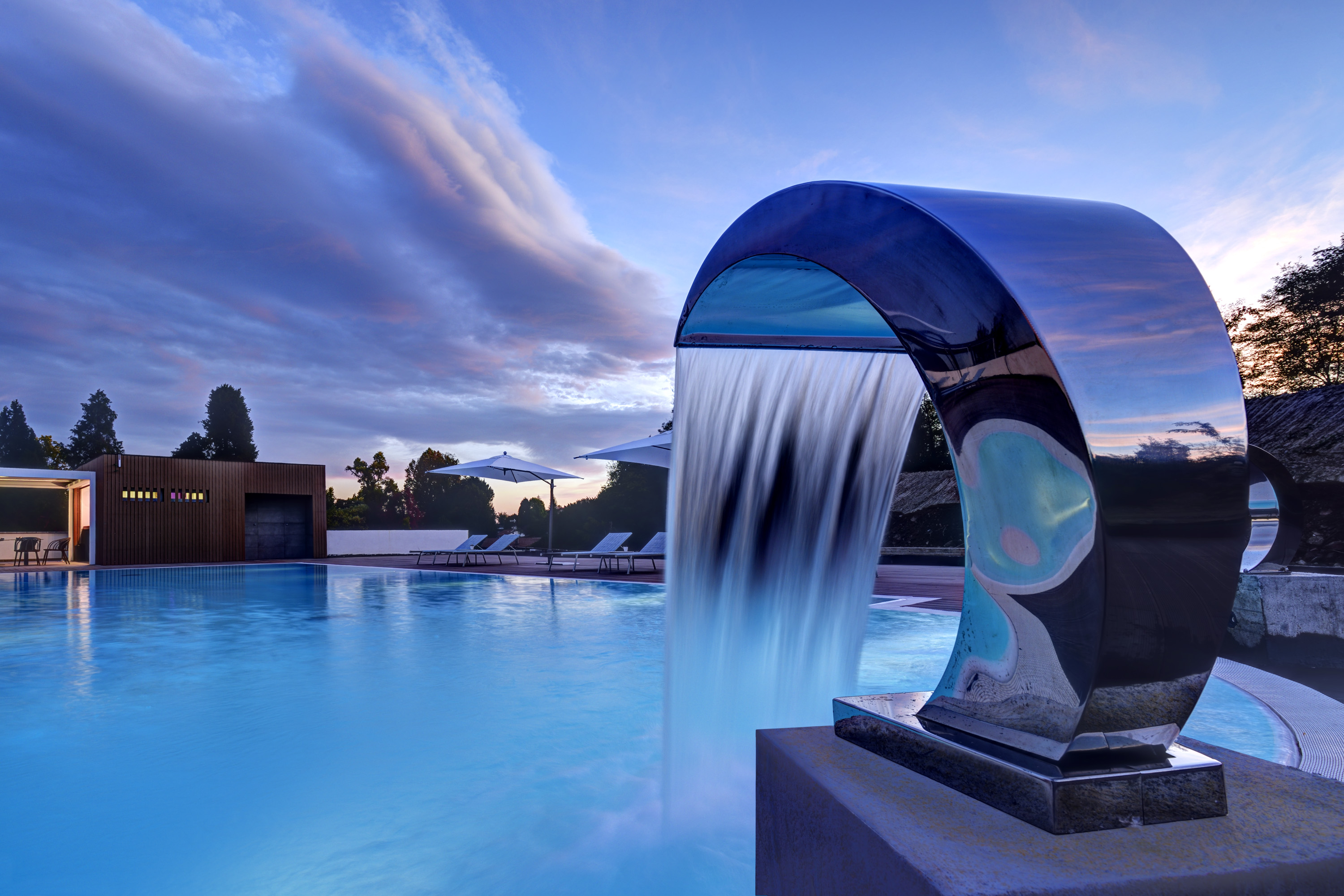 A curved metallic waterfall feature cascading water into a resort swimming pool at dusk, with dramatic purple and blue clouds filling the sunset sky.