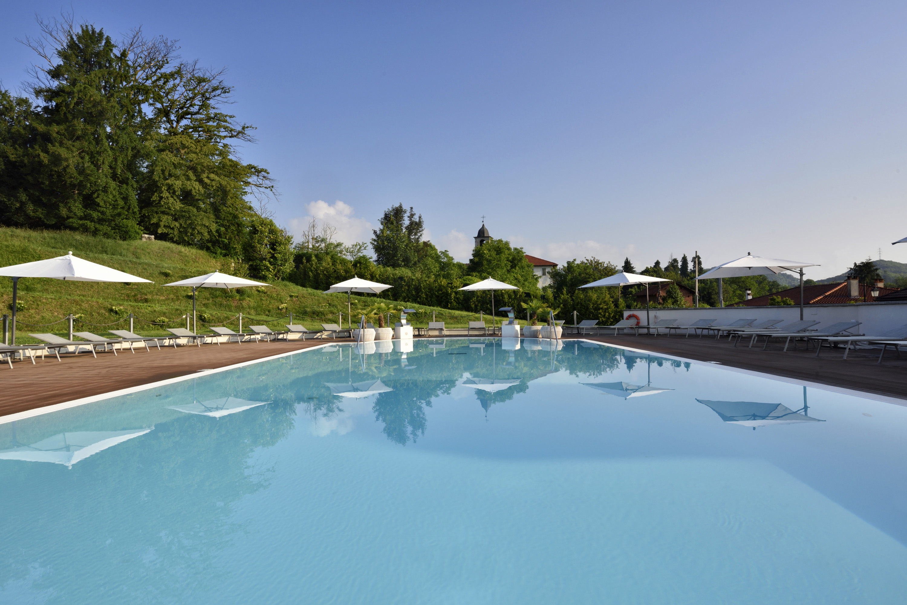 A serene outdoor swimming pool reflecting white patio umbrellas and lush green trees along a grassy hillside, with lounge chairs lining the wooden deck under a clear blue sky.