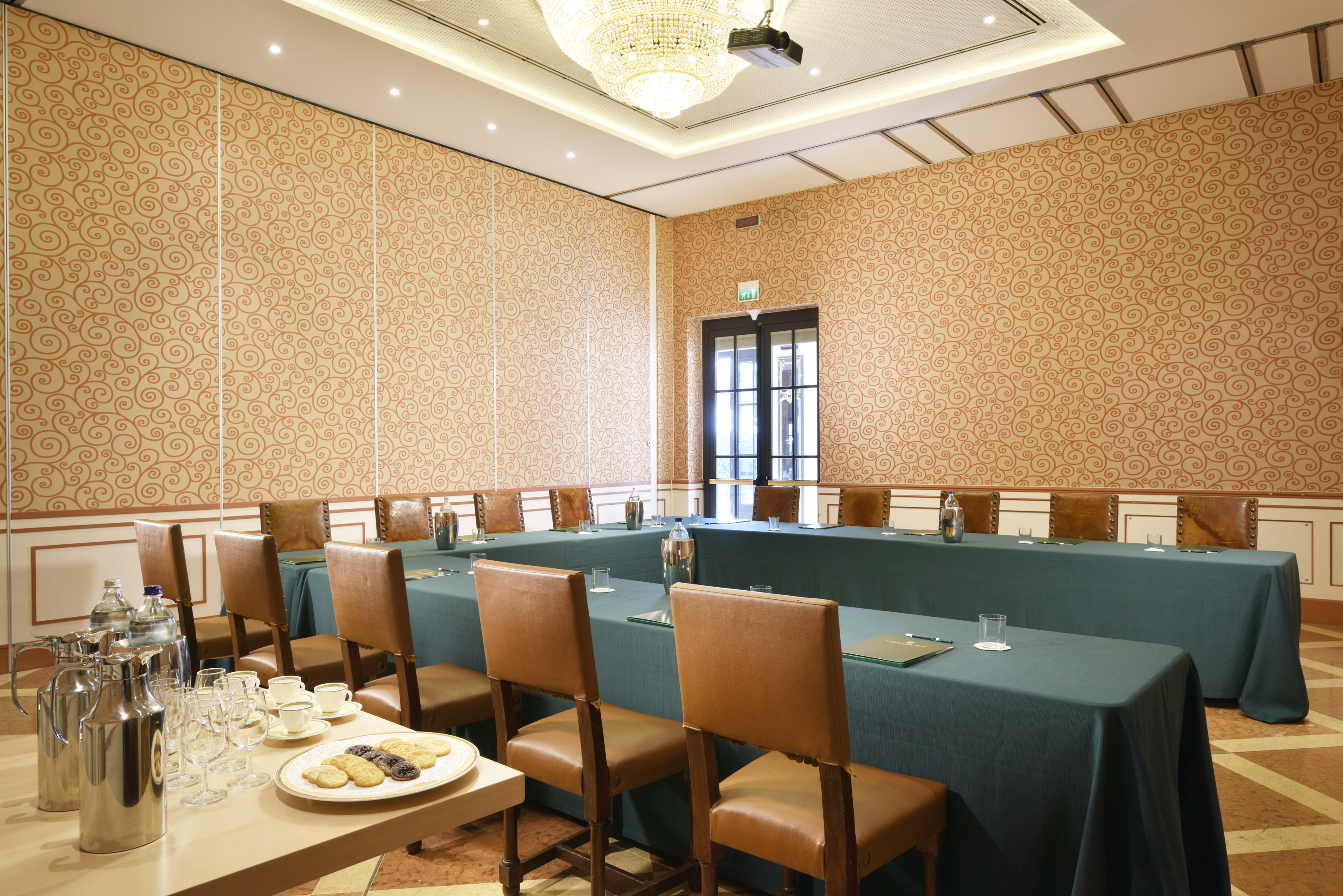 A formal meeting room configured in a U-shape layout with teal table linens, cognac leather chairs, patterned wallpaper, a crystal chandelier, and a refreshment table in the foreground.