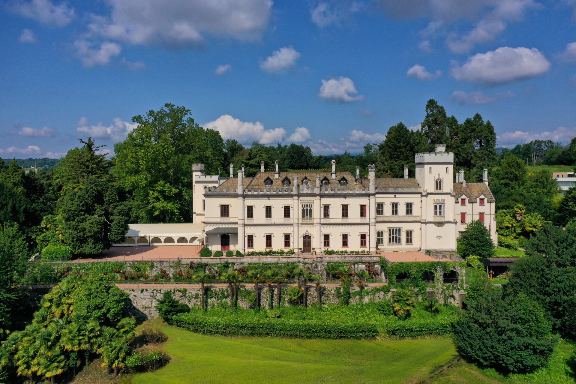 A wide aerial view showcasing the front facade of a majestic white historic estate with crenellated towers, terraced gardens, and lush green lawns under a blue sky.