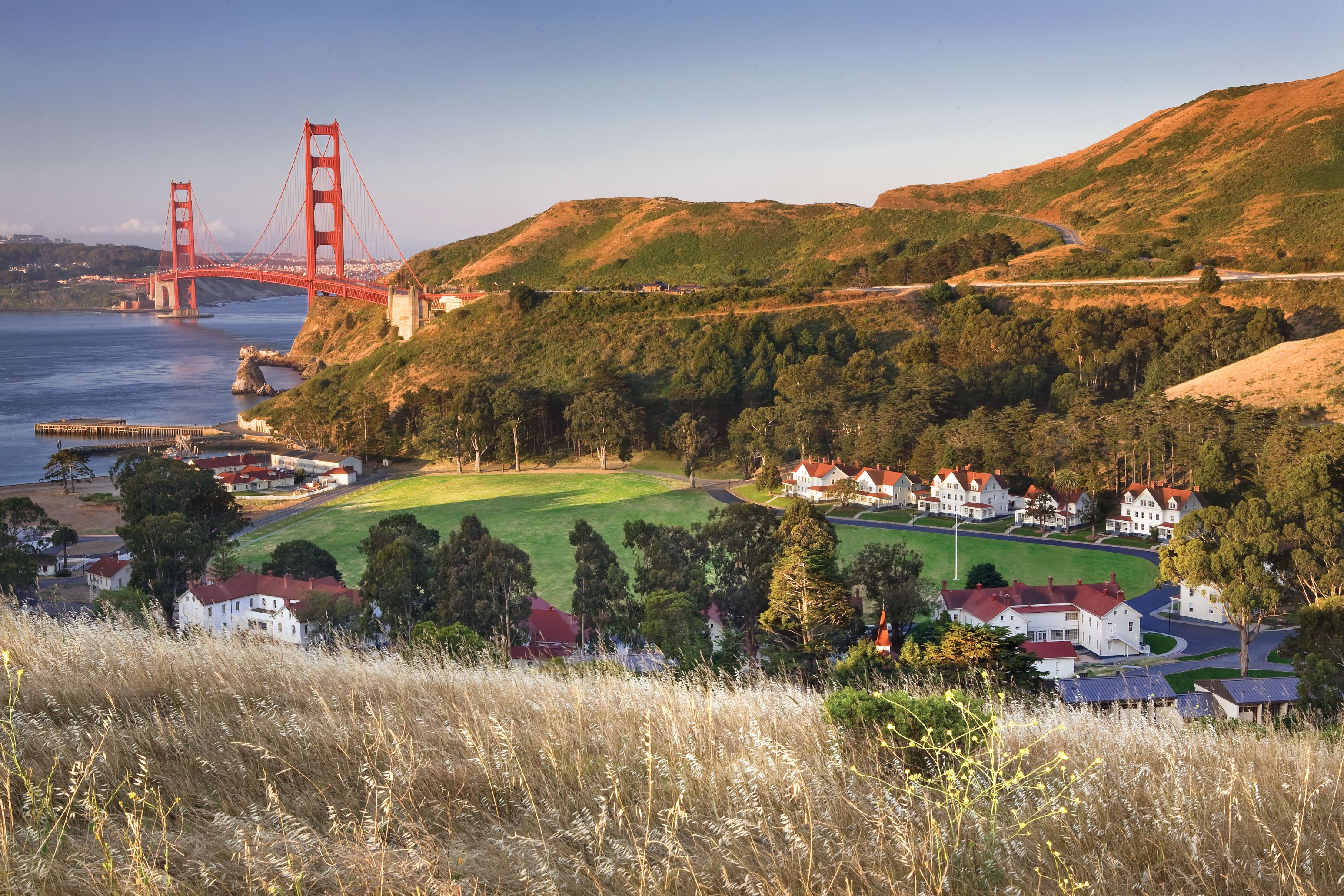 A scenic elevated view of Cavallo Point Lodge at Fort Baker nestled in coastal green hills, with red-roofed historic white buildings clustered around a central parade lawn and the Golden Gate Bridge visible in the background.