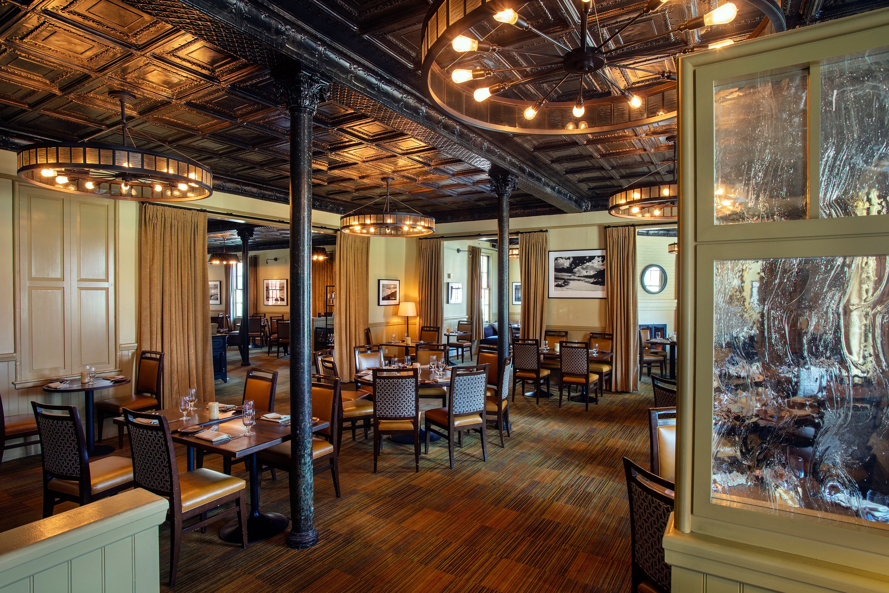 A warm, atmospheric restaurant space with a pressed-tin ceiling, round wagon-wheel chandeliers, dark support columns, beige drapes, and wooden tables with pattern-backed dining chairs.