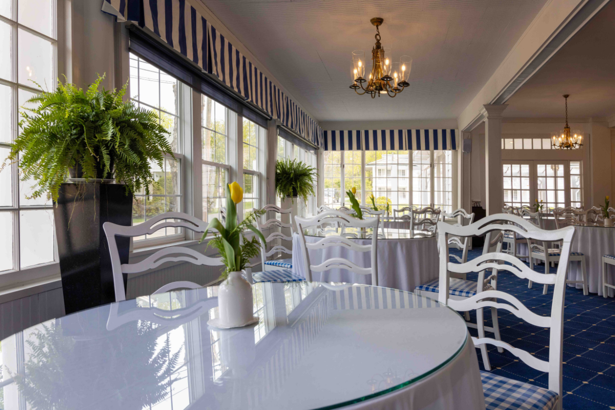 Bright coastal-style dining room featuring large sunlit windows, white wooden chairs with blue gingham seat cushions, glass-topped tables, blue-and-white striped valances, and brass chandeliers.