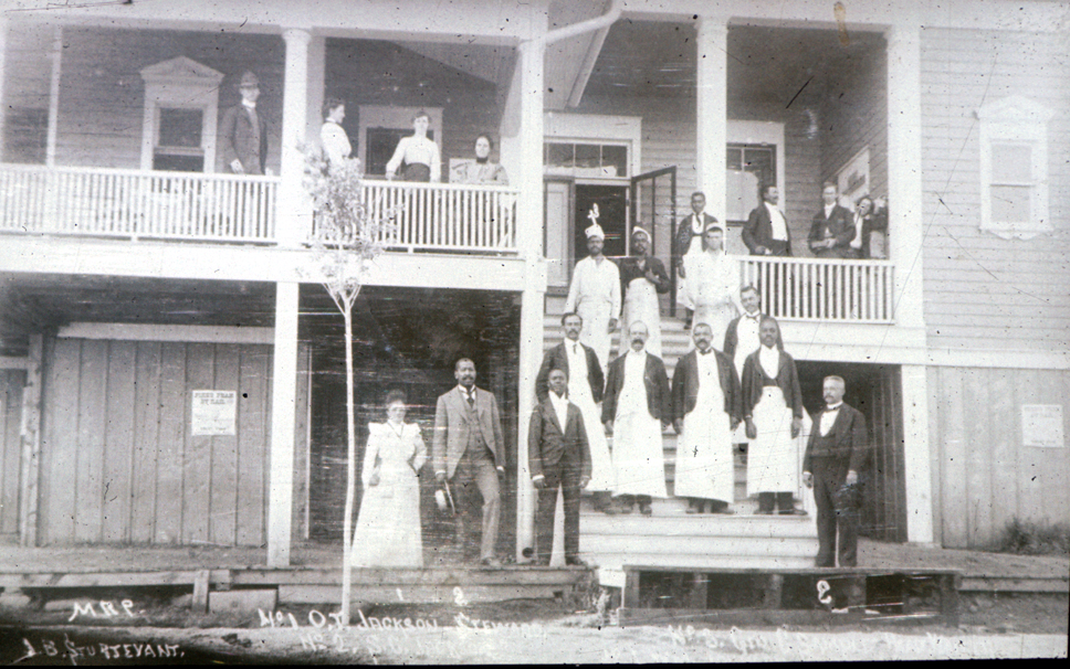 A historical black-and-white photograph of hospital or hotel staff posing outside a two-story wooden building with a porch, including servers, cooks, and managers.