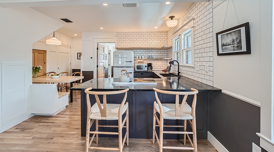 A modern open-plan suite kitchen featuring a black-lacquered island with wishbone bar stools, dark countertops, white subway tiled walls, and an adjacent light wood dining nook.