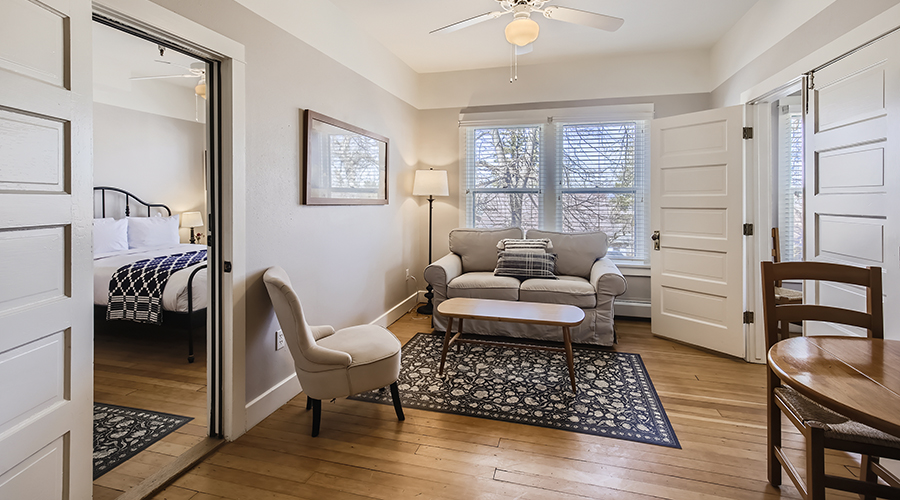 A warm, cozy hotel suite featuring light-colored walls, a small seating area with a sofa and accent chair on an area rug, and an open door leading to an adjoining bedroom.