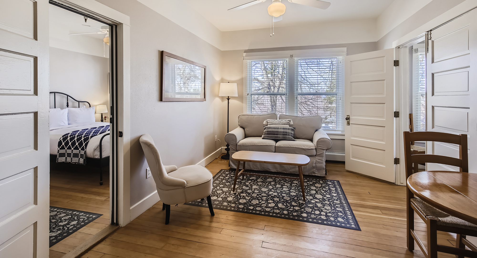 A bright, minimalist hotel suite living room with light hardwood floors, a neutral beige sofa, an accent armchair, a patterned area rug, and an adjoining doorway showing a glimpses of a wrought-iron bed.