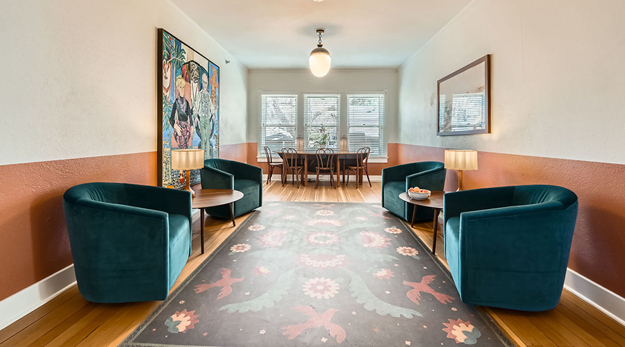 A stylish lounge room featuring deep teal velvet armchairs facing each other on a patterned rug, terracotta-and-white dual-tone walls, a large colorful painting, and a small dining nook by the window.