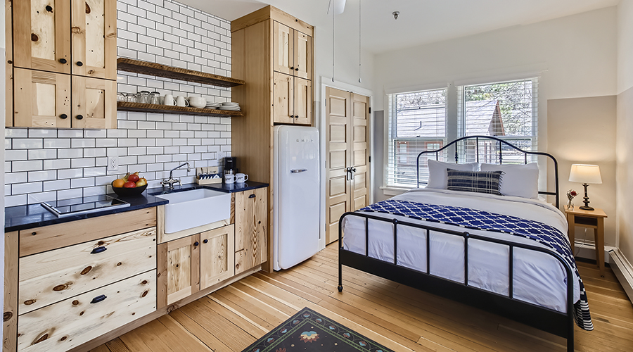 A rustic studio suite combining a fully equipped kitchen with natural pine cabinets, white subway tile backsplashes, and a Smeg refrigerator, alongside a cozy sleeping area featuring a black wrought-iron bed dressed in blue and white linens.