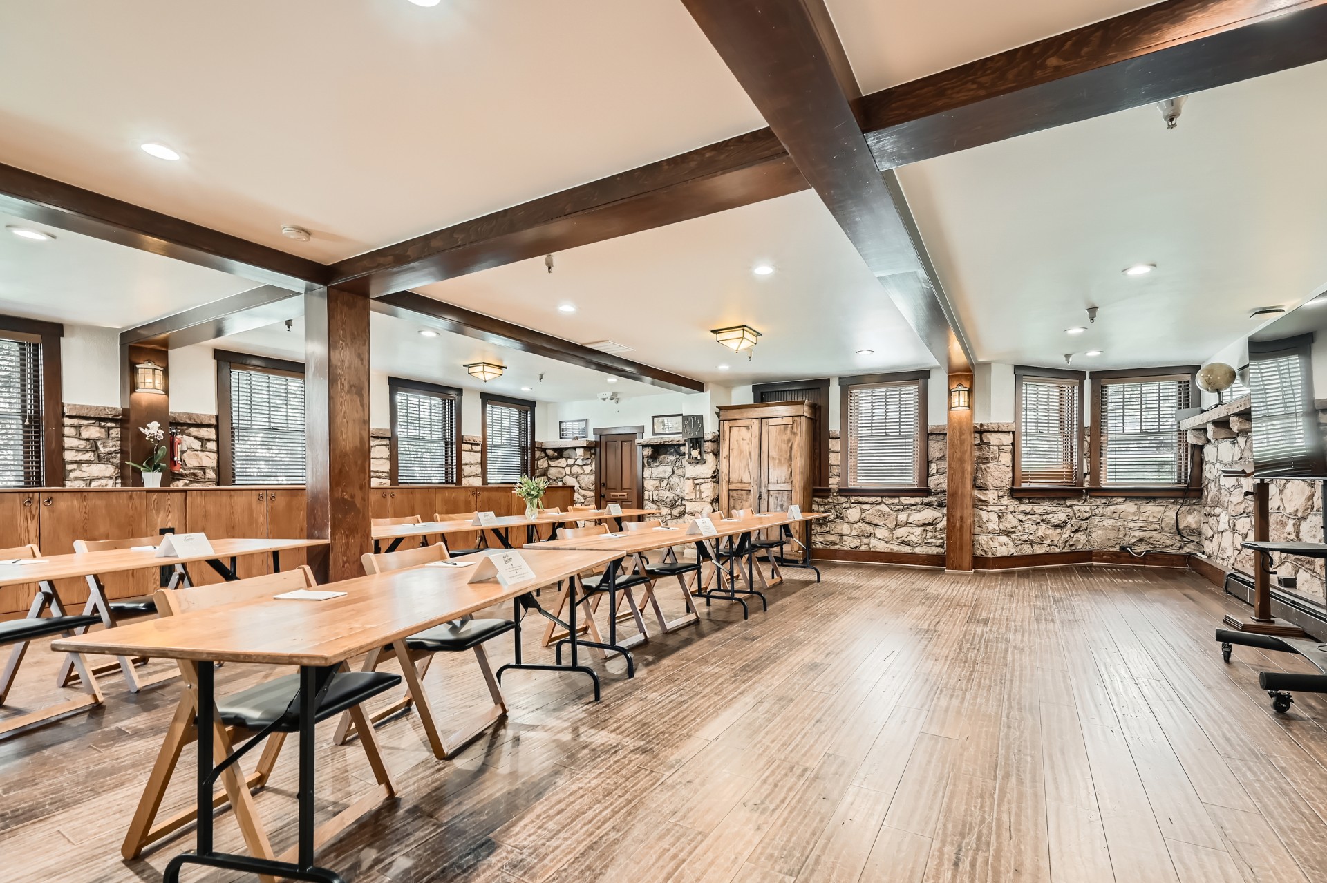 A rustic classroom-style meeting space with exposed wooden ceiling beams, stone accent walls, hardwood flooring, and long wooden tables facing a presentation area.